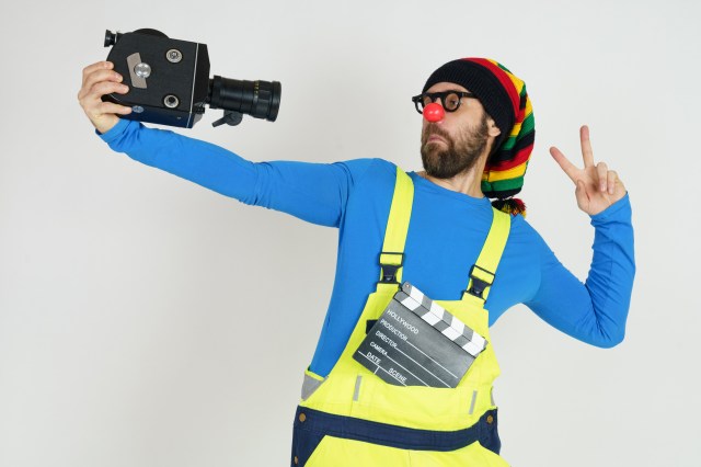 Celebration and communication concept. A clown in bright clothes filming himself with a movie camera. Isolated on white