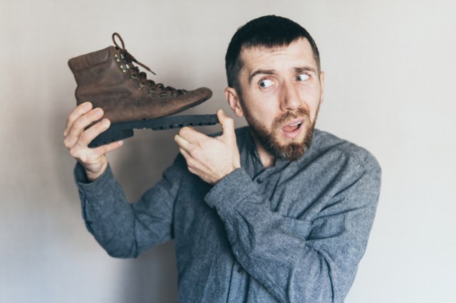 Young bearded Caucasian man holds old leather boot with torn sole, listening, surprise and shock on his face